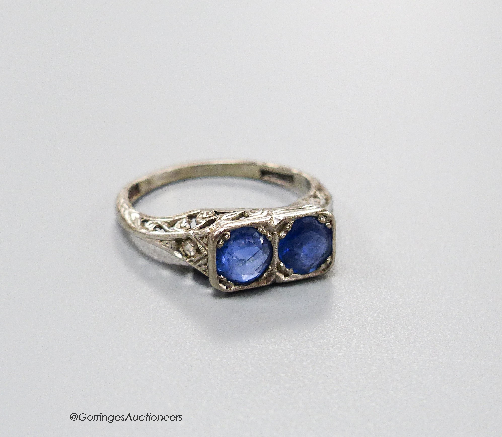 A 1920's white metal (stamped 18ct & plat) and two stone sapphire set ring with carved and single stone diamond chip set shoulders, size K, gross weight 3.2 grams